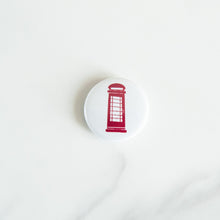 Load image into Gallery viewer, A button on white tabletop. The button features an illustration of a red telephone booth. 