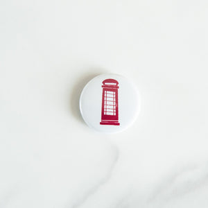 A button on white tabletop. The button features an illustration of a red telephone booth. 