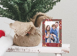 A photo of a one-sided Christmas card showing the front of the card standing up next to the bottom of a small Christmas tree with the base wrapped in burlap. The photo card features one photo with a frame of illustrated leaves in various colors. Below the photo it reads “Rejoice Greatly” and has space to put your family name. 
