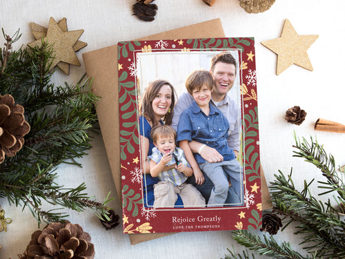 A photo of a one-sided Christmas card showing the front of the card on top of a brown wrapped gift on a white tabletop. Around the gift are pine needles, pinecones and wood star ornaments. The photo card features one photo with a frame of illustrated leaves in various colors. Below the photo it reads “Rejoice Greatly” and has space to put your family name. 