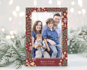 A photo of a one-sided Christmas card showing the front of the card standing up with pine needles behind and blurred white Christmas lights. The photo card features one photo with a frame of illustrated leaves in various colors. Below the photo it reads “Rejoice Greatly” and has space to put your family name. 