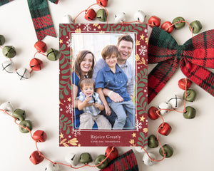 A photo of a one-sided Christmas card showing the front of the card laying on white table top. The card is surrounded by plaid red and green ribbon and red, green and white bells. The photo card features one photo with a frame of illustrated leaves in various colors. Below the photo it reads “Rejoice Greatly” and has space to put your family name. 