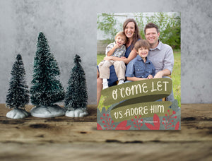 A photo of a one-sided Christmas card showing the front of the card standing up with three small Christmas trees next to it. The photo card features one photo with the words “O Come Let Us Adore Him” below with illustrated red leaves.