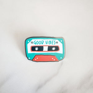 An enamel pin featuring the words "good vibes" inside an illustrated cassette tape.