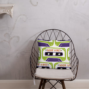 A white pillow on a metal chair. The white pillow features the artwork on a white background with an illustrated cassette tape pattern with the words 'good vibes.'