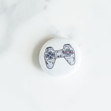 Load image into Gallery viewer, A button pin on a white tabletop. The button features an illustration of a retro Playstation-like game controller. 