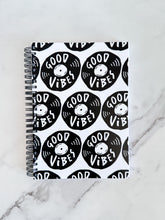 Load image into Gallery viewer, A spiral notebook is on a tabletop. The white spiral notebook has black spiral binding. The pattern design features the words &quot;good vibes&quot; inside an illustrated vinyl record.  