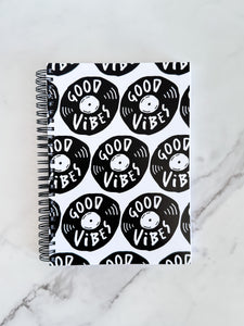 A spiral notebook is on a tabletop. The white spiral notebook has black spiral binding. The pattern design features the words "good vibes" inside an illustrated vinyl record.  