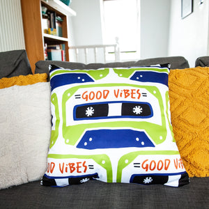 A white pillow on a grey sofa with other pillows. The white pillow features the artwork on a white background with an illustrated cassette tape pattern with the words 'good vibes.'