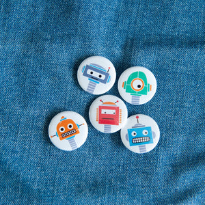 A set of five button pins on a jean jacket. The buttons feature five different illustrations of robots.