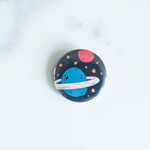 Load image into Gallery viewer, A button on a white tabletop. The button features space-themed illustration of planet. 