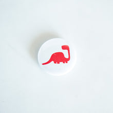 Load image into Gallery viewer, A button pin on a white tabletop. The button features an illustration of a Sauropod. 