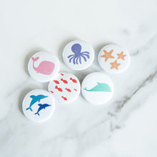 Load image into Gallery viewer, A set of six buttons on a white tabletop. The buttons feature illustrations of whales, fish, starish, dolphins, and an octopus. 