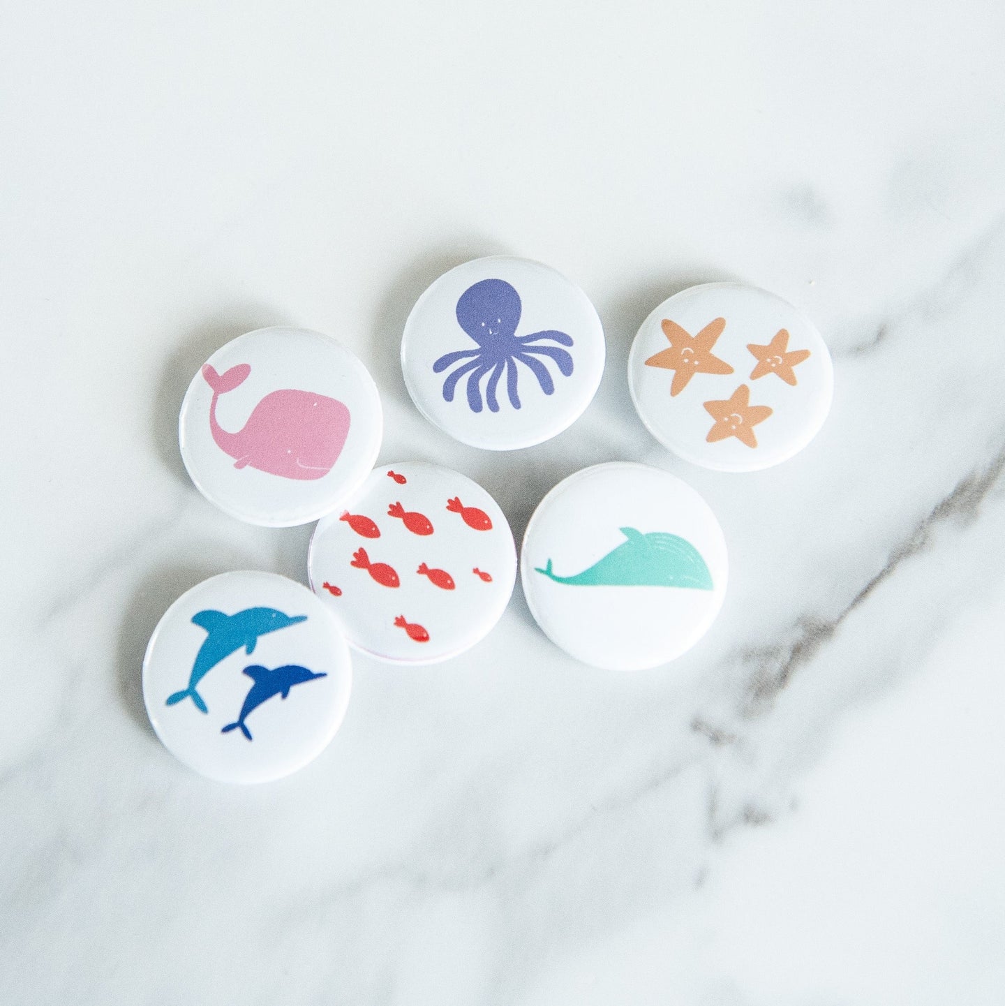 A set of six buttons on a white tabletop. The buttons feature illustrations of whales, fish, starish, dolphins, and an octopus. 