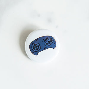 A button pin on a white tabletop. The button features an illustration of a retro Sega Genesis-like game controller. 