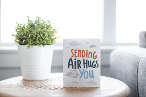 A greeting card laying on a wooden table with some cut wood details. The card features the words “Sending air hugs to you.”