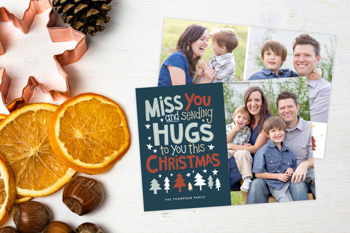 A photo of a Christmas card showing the front and back of the card laying on a white surface. Left of the card is a cookie cutter, pinecone, nuts and dried oranges. The front of the card features a photo on the right side and on the left side are the words “ Miss you and sending hugs to you this Christmas” with illustrated trees below the words. The back of the card features two photos with illustrated trees at the bottom.