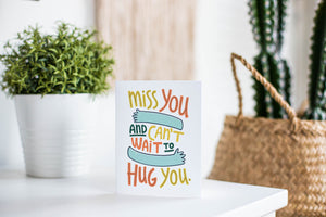 A greeting card is featured on a white tabletop with a white planter in the background with a green plant. There’s a woven basket in the background with a cactus inside. The card features the words “Miss you and can’t wait to hug you.”