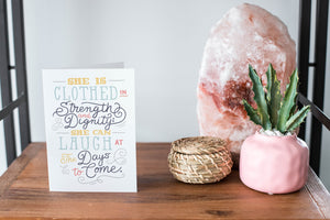 A card on a wood tabletop and on the right side of the card is a woven basket, a pink plant pot with a cactus in it and a pink crystal rock. The card features the words "She is clothed in strength and dignity; she can laugh at the days to come."