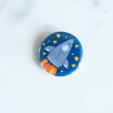 Load image into Gallery viewer, A button on a white tabletop. The button features space-themed illustration of a rocket ship. 