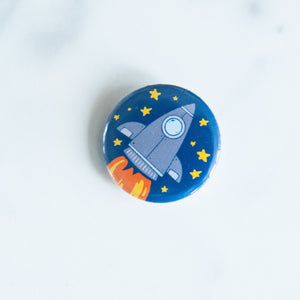 A button on a white tabletop. The button features space-themed illustration of a rocket ship. 