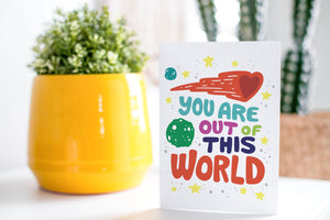 A greeting card is on a table top with a yellow plant pot and a green plant inside. The card features the words "You are out of this world” with space themed illustrations.