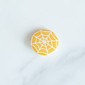 A button on a white tabletop. The button features an illustration of a spiderweb.