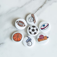 Load image into Gallery viewer, A set of buttons on a white tabletop. The buttons feature illustrations of an American football, a soccer/football, a baseball with baseball bat, a rugby ball, a cricket ball and bat, a basketball and a golf club with ball. 