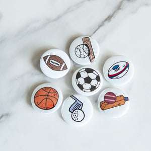 A set of buttons on a white tabletop. The buttons feature illustrations of an American football, a soccer/football, a baseball with baseball bat, a rugby ball, a cricket ball and bat, a basketball and a golf club with ball. 