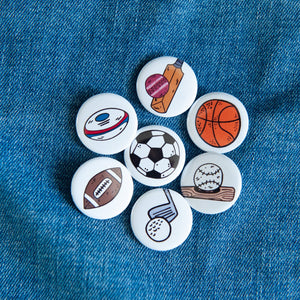 A set of buttons on a jean jacket. The buttons feature illustrations of an American football, a soccer/football, a baseball with baseball bat, a rugby ball, a cricket ball and bat, a basketball and a golf club with ball. 