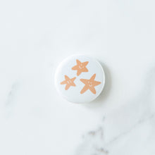 Load image into Gallery viewer, A button on a white tabletop. The button features an illustration of starish. 