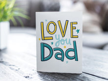 Load image into Gallery viewer, A photo of a card featured on a tabletop next to a white planter filled with a green plant. ​​The card features the words “Love you Dad” with small stars around the letters.