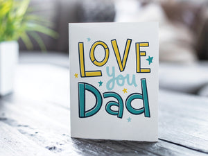A photo of a card featured on a tabletop next to a white planter filled with a green plant. ​​The card features the words “Love you Dad” with small stars around the letters.