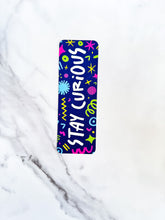 Load image into Gallery viewer, A bookmark placed on a tabletop. The bookmark has the words &quot;stay curious&quot; with fun, sketchy illustrations. 