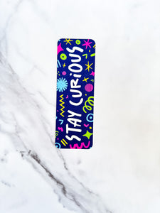 A bookmark placed on a tabletop. The bookmark has the words "stay curious" with fun, sketchy illustrations. 