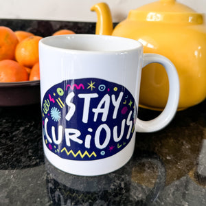 A white mug on a black countertop with a tea pot and oranges behind it. The mug features a design with the words 'stay curious' in an illustrated oval shape with sketchy elements around it. 