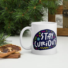 Load image into Gallery viewer, A white mug on a tabletop with part of a pine tree in the background. The mug features a design with the words &#39;stay curious&#39; in an illustrated oval shape with sketchy elements around it. 