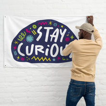 Load image into Gallery viewer, A man hanging a large white flag on a wall. The white flag features the words &quot;stay curious&quot; featured inside a a purple shape with sketchy illustrations around. 