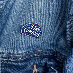 An enamel pin on a jacket with the words "stay curious" surrounded by whimsical sketches. 