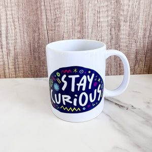 A white mug on a tabletop. The mug features a design with the words 'stay curious' in an illustrated oval shape with sketchy elements around it. 