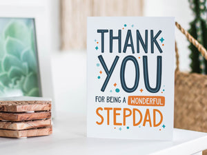 A greeting card is featured on a white tabletop with a white planter in the background with a green plant. There’s a woven basket in the background with a cactus inside. The card features the words “Thank You for Being a Wonderful Stepdad.”
