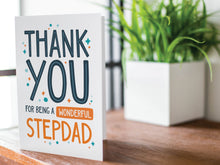 Load image into Gallery viewer, A greeting card is featured on a wood coffee table with a green plant in a white planter in the background. The card features the words “Thank You for Being a Wonderful Stepdad.” 