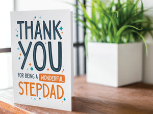 A greeting card is featured on a wood coffee table with a green plant in a white planter in the background. The card features the words “Thank You for Being a Wonderful Stepdad.” 