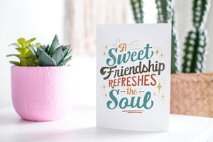 A greeting card featured standing up on a white tabletop with a pink plant pot in the background and some succulents in the pot. There’s a woven basket in the background with a cactus inside. The card features the words “A sweet friendship refreshes the soul.”  