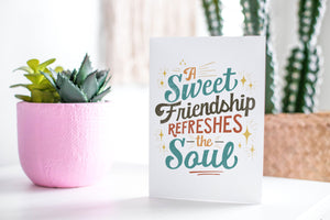 A white greeting card sitting on a a white table top with plants in the background. The card has the words "A Sweet Friendship Refreshes the Soul."