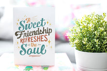 Load image into Gallery viewer, A greeting card is on a table top with a gift in pink wrapping paper. Next to the gift is a white plant pot with a green plant. The card features the words “A sweet friendship refreshes the soul.”