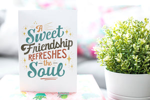 A greeting card is on a table top with a gift in pink wrapping paper. Next to the gift is a white plant pot with a green plant. The card features the words “A sweet friendship refreshes the soul.”