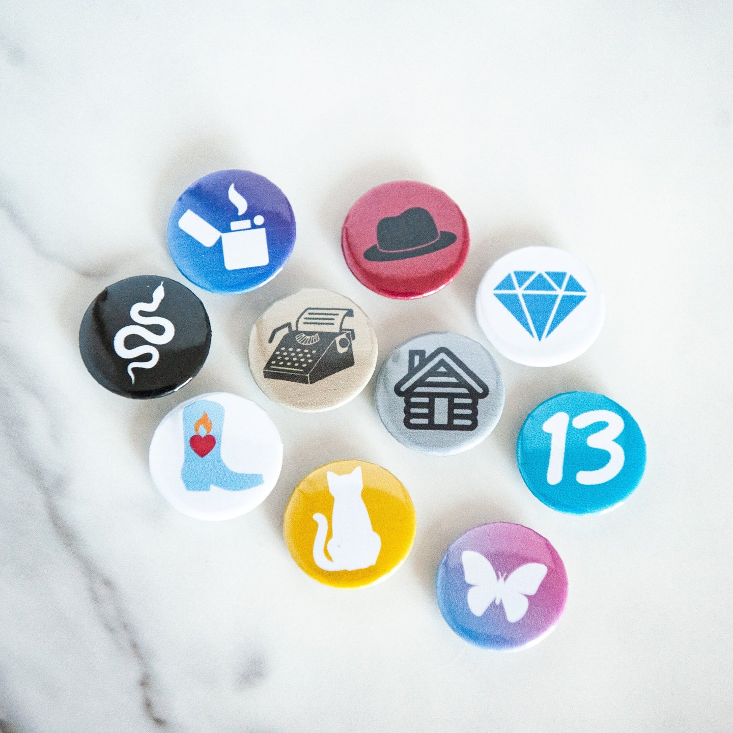 A photo of 10 pin badges on a white tabletop. The badges feature illustrations of a snake, a lighter, a cowboy boot,  cat, a butterfly, a house, a typewriter, a diamond, the number 13, and a black hat. 