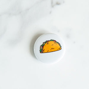 A button pin on a white tabletop. The pin features an illustration of a taco.