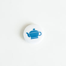 Load image into Gallery viewer, A button on white tabletop. The button features an illustration of a blue teapot. 